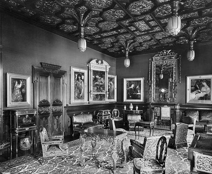 A drawing room in 1892 with framed pictures hung at eye level around a carved cabinet, a mirror and a table